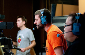 A male student wears a headset and looks through a video camera