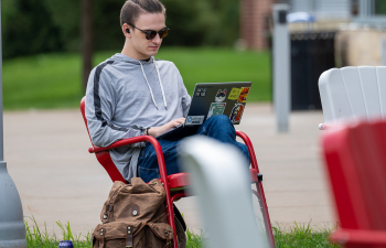 A male student sits outside the University Center doing homework on their laptop