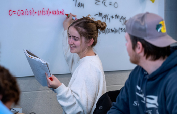 A female student writes a math equation on a whiteboard in class