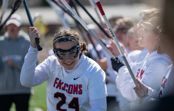 a female lacrosse player wearing a white uniform and black goggles runs through an arch of her teammates' sticks