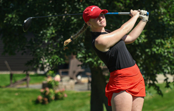 a female golfer wearing a black polo, red skirt and red cap swings a golf club