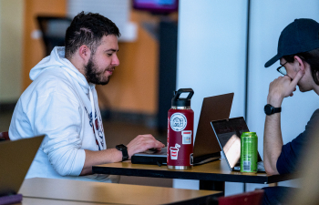 Two male students sit at a table in the University Center with their laptops