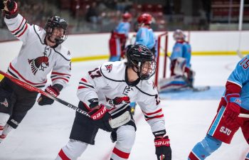 two UWRF hockey players in white jerseys, black helmets and black hockey pants celebrate a goal while players in blue uniforms behind them look defeated