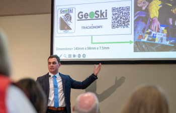 a male presenter stands in front a power point projected on a screen