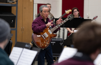 a faculty member holding a guitar speaks to students sitting in chairs with music stands in front of them