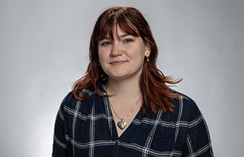 headshot of a white female. She has long red hair and is wearing a dark plaid shirt.