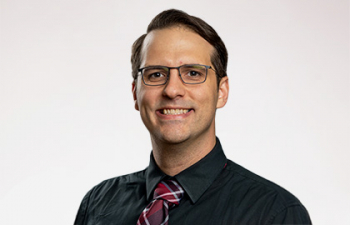 Headshot of Scott Mitchell, a man with glasses, brown hair and a black shirt with a red tie