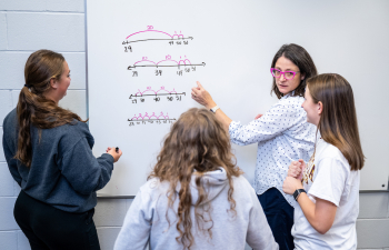 a teacher stands at a whiteboard and talks with a group of three female students