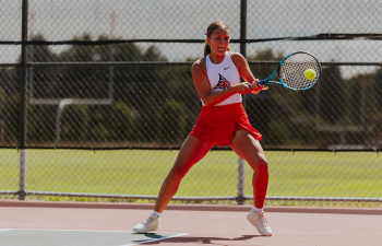 a women's tennis player wearing a white top and red tennis skirt swings a tennis racket at a tennis ball