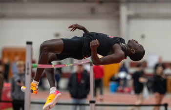 a Black man wearing a black track uniform jumps over the high jump bar 