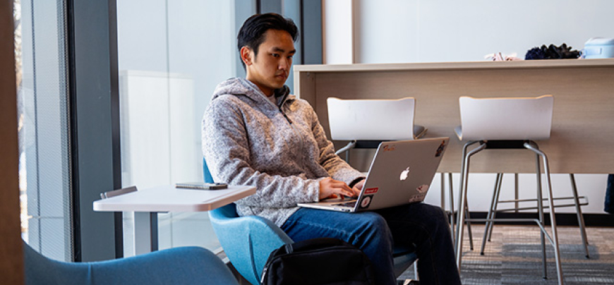 A male student sits in a SciTech commons space on their laptop